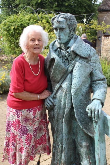 A smiling elderly woman in a red top and floral skirt stands next to a bronze statue of a man in a coat, set in a garden with greenery in the background.