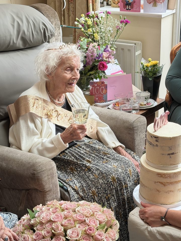 An elderly woman wearing a tiara and a sash is seated in an armchair, holding a glass of champagne. She is surrounded by flowers, birthday cards, and a tiered cake. People are gathered around her, celebrating her 100th birthday.